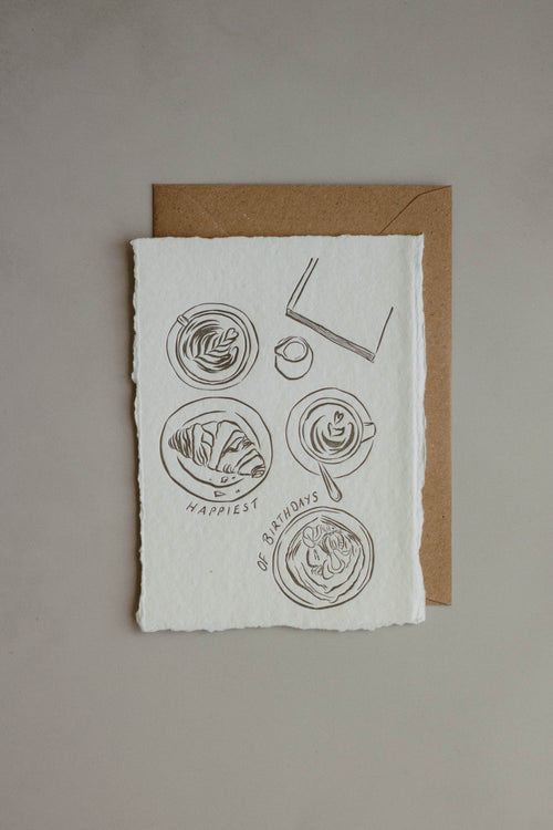 An A6 handmade paper card with rough edges, featuring a minimalist charcol ink illustration of coffee cups and a book, with the text 'Happiest of Birthdays' and displayed on top of a brown kraft envelope against a neutral grey background.