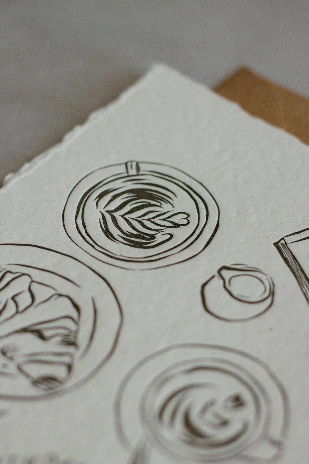 A close-up of textured off-white handmade paper featuring hand-drawn charcol ink sketches of coffee which has been letterpresed.