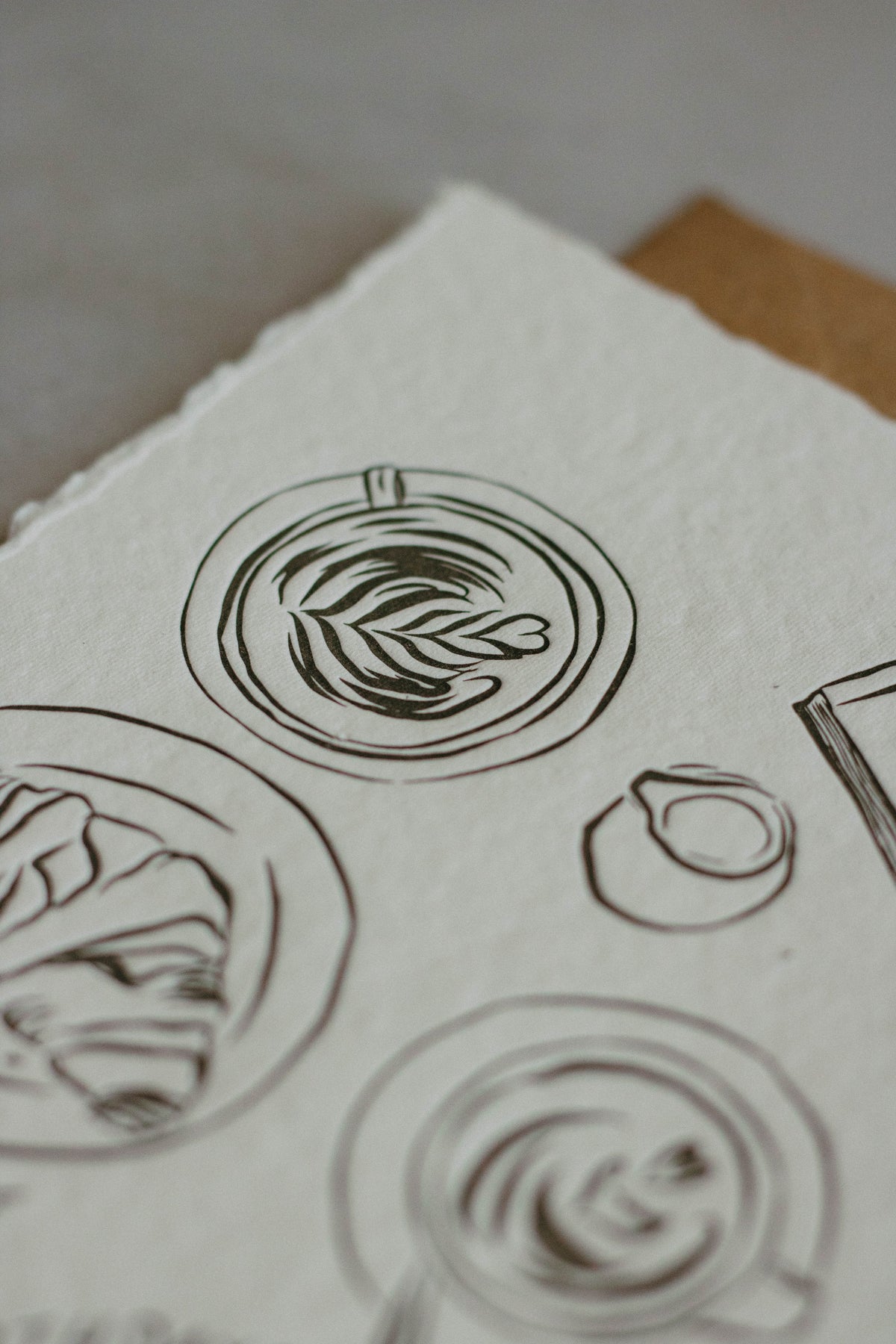 A close-up of textured off-white handmade paper featuring hand-drawn charcol ink sketches of coffee which has been letterpresed.