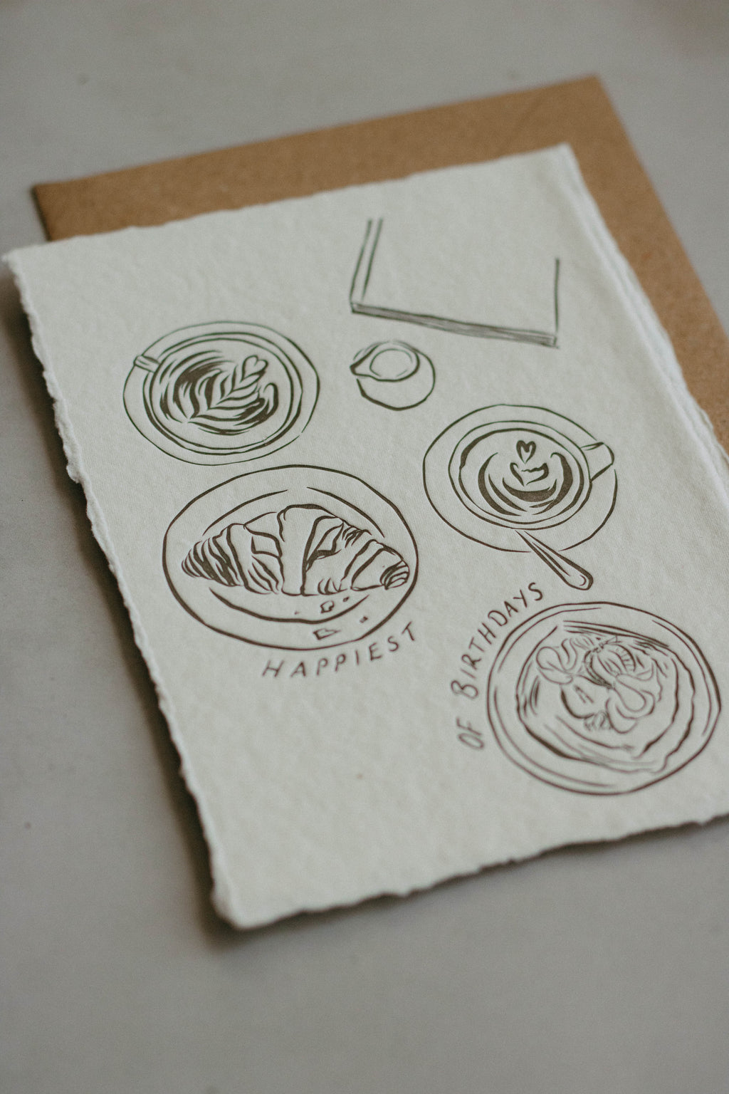 An A6 artwork with rough, deckled-edge white paper featuring minimalist charcol line drawings of coffee cups, a croissant, and a coffee with the text 'HAPPIEST OF BIRTHDAYS' printed below, displayed on a brown kraft envelope against a neutral grey background.