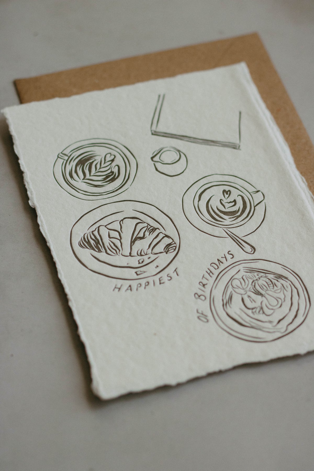 An A6 artwork with rough, deckled-edge white paper featuring minimalist charcol line drawings of coffee cups, a croissant, and a coffee with the text 'HAPPIEST OF BIRTHDAYS' printed below, displayed on a brown kraft envelope against a neutral grey background.