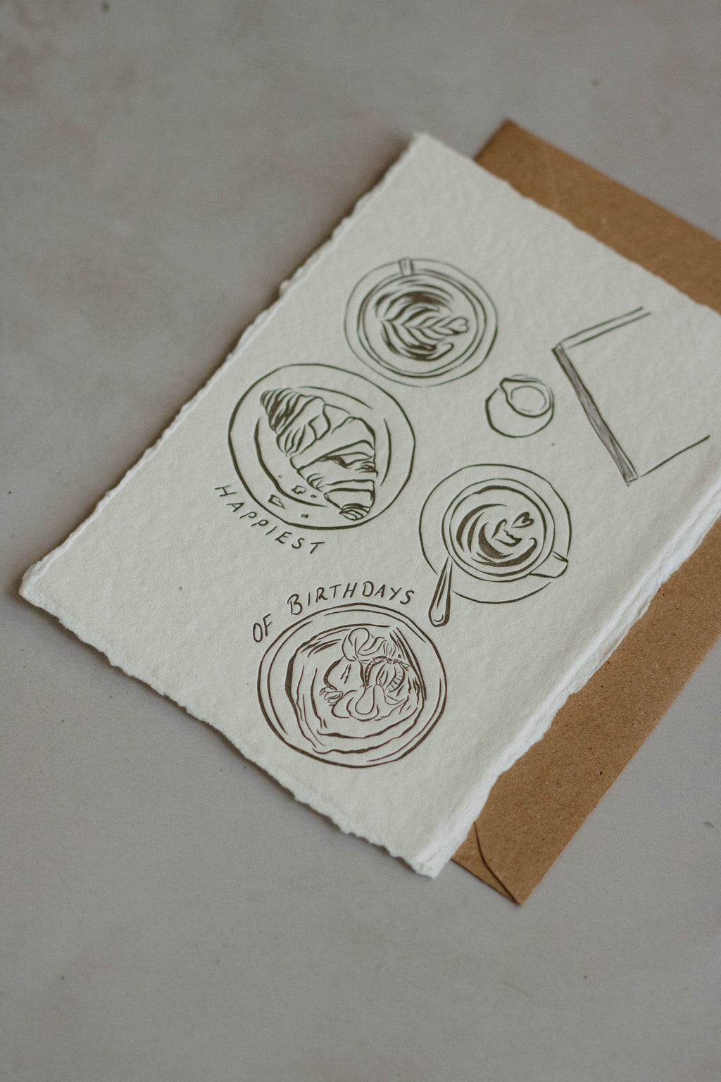 A6 handmade paper birthday note card with rough deckled edges, featuring minimalist charcol ink drawings of coffees and pastries and the text 'HAPPIEST OF BIRTHDAYS', placed on top of a brown kraft envelope against a neutral background.