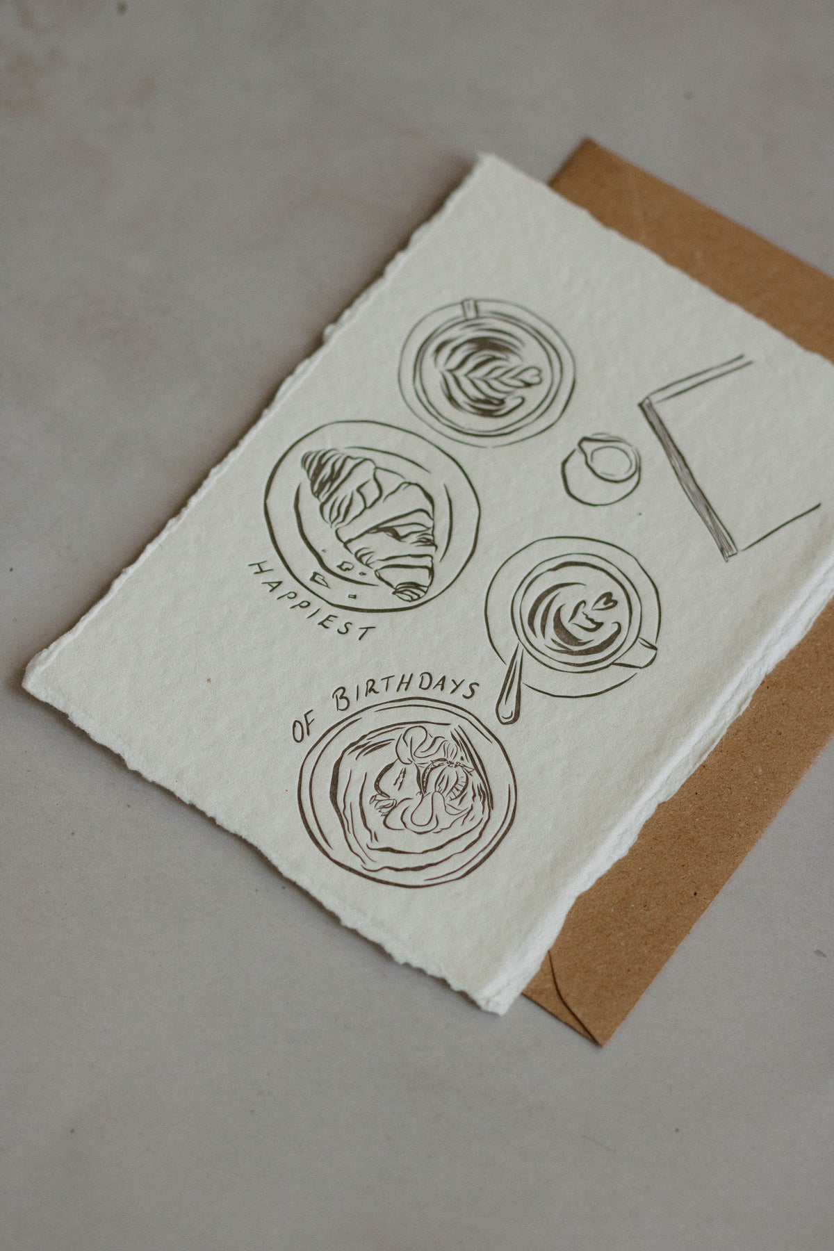 A6 handmade paper birthday note card with rough deckled edges, featuring minimalist charcol ink drawings of coffees and pastries and the text 'HAPPIEST OF BIRTHDAYS', placed on top of a brown kraft envelope against a neutral background.
