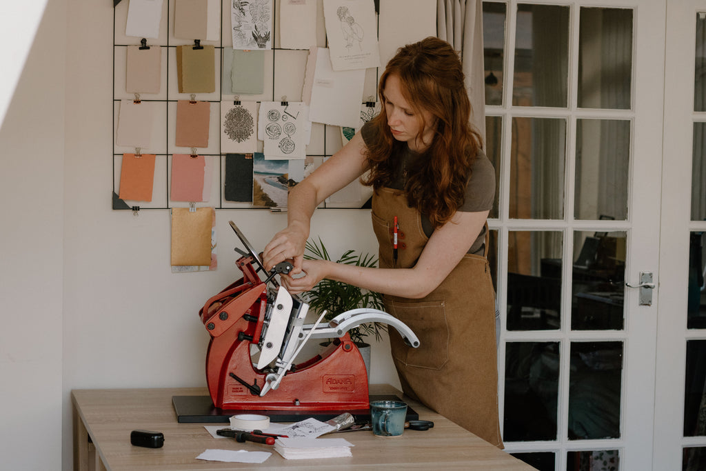 A woman in a brown apron stands at a table using a red and silver traditional letterpress printer, with sketches pinned on a board.