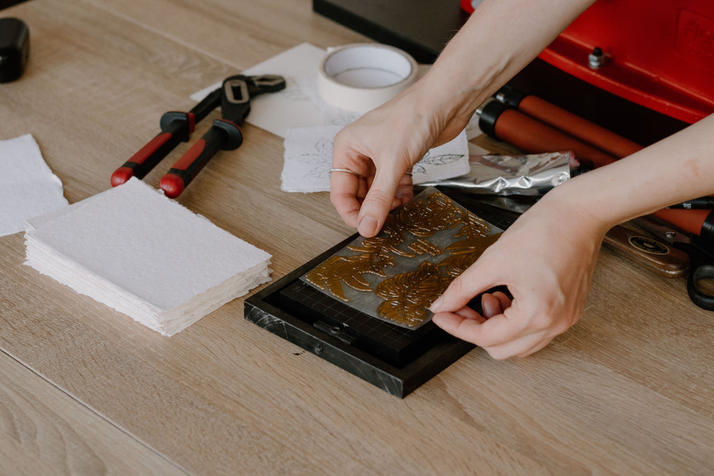 A close-up of a person's hands applying a designed plate onto a black metal block ready for printing, set on a light wood desk surrounded by crafting tools and natural handmade paper.